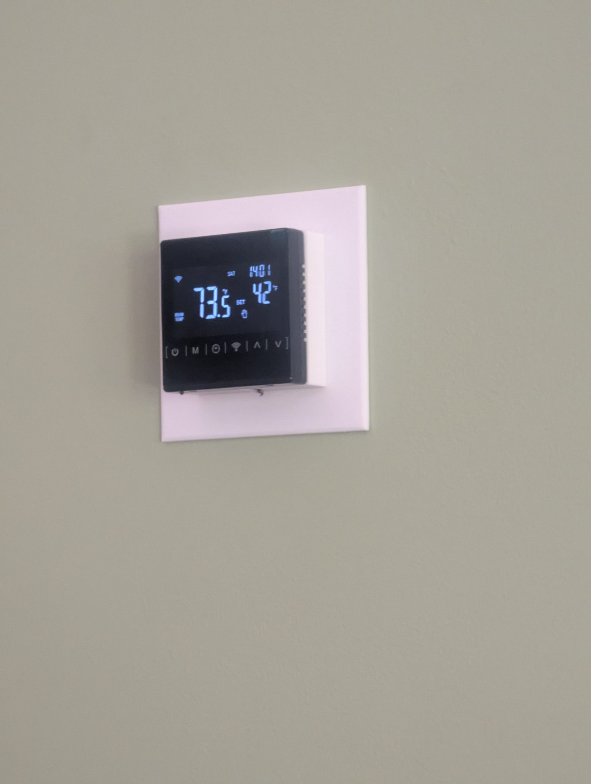 A photograph of a green wall with a black digital thermostat and a white square around the thermostat. The screen reads 73.5° Fahrenheit. with the set point of 42° Fahrenheit